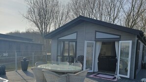 Une terrasse extérieure moderne, dotée d’une porte vitrée donnant sur un patio abrité, entourée d’une terrasse en bois et d’un jardin clôturé.