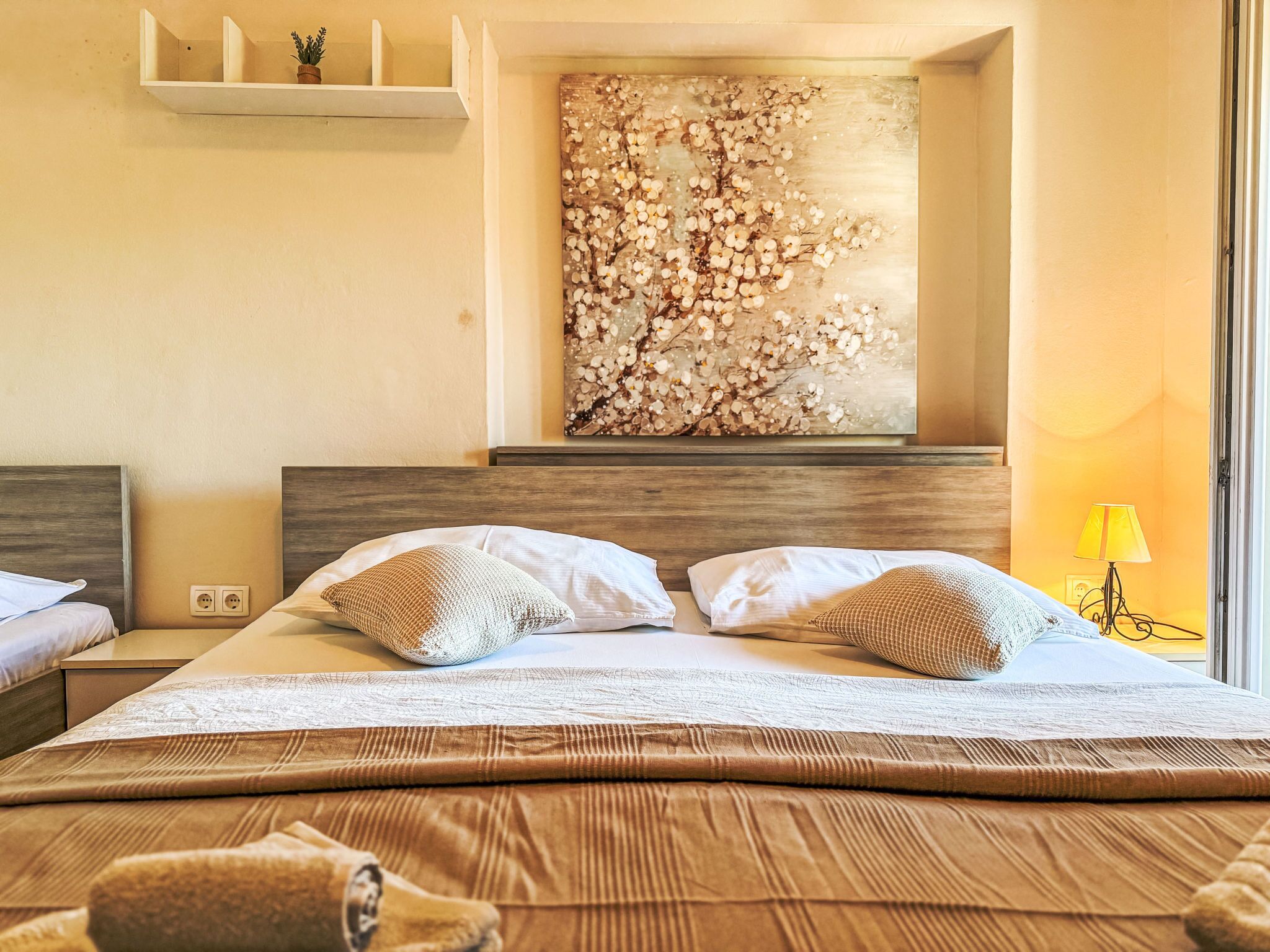 A hotel room with a large bed, a wooden headboard, a wall-mounted shelf with a potted plant, and a large floral artwork above the bed.