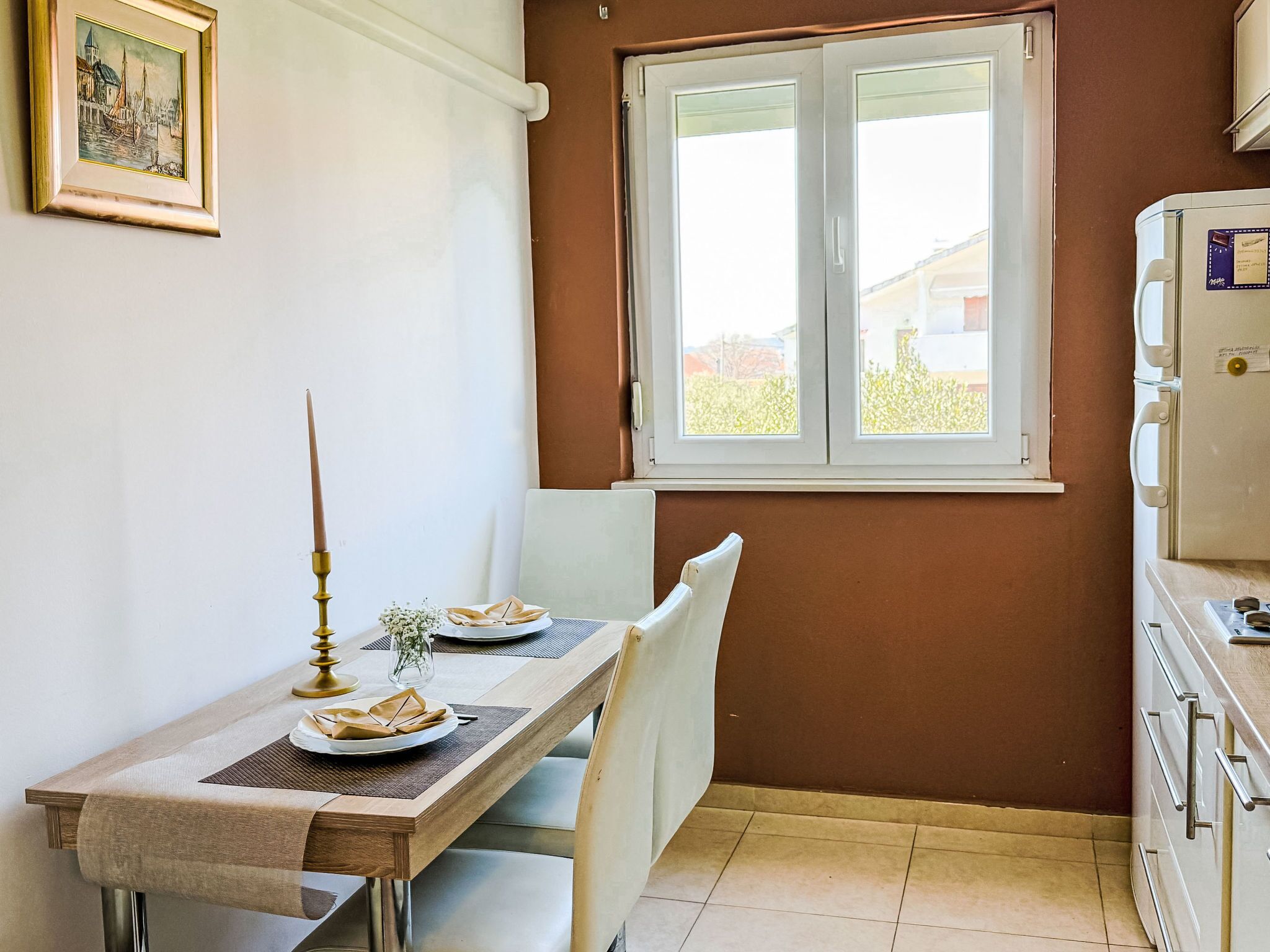 A small kitchen with a dining table set for two, a window, and a refrigerator.