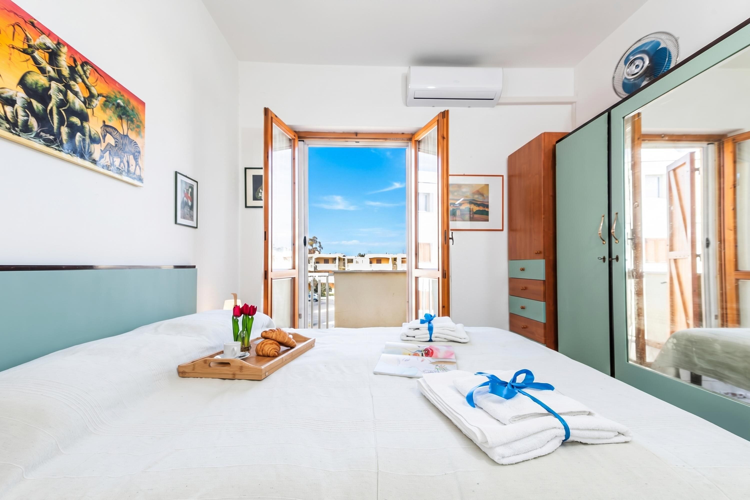 A hotel room with a bed, a breakfast tray, towels, a painting, and a view of the outside.