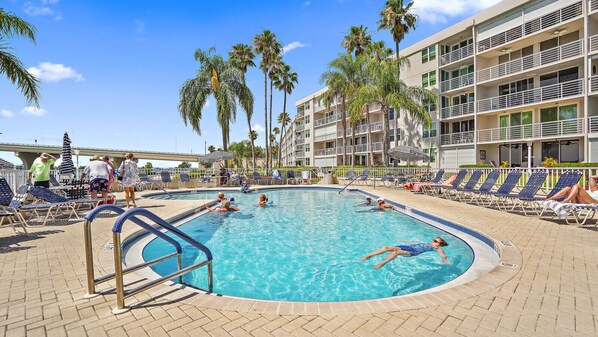 Una piscina rodeada de sillas de descanso y palmeras, con un edificio de varios pisos al fondo.