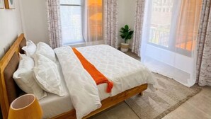A neatly made bed with white linens and an orange throw, a wooden nightstand with a lamp, and a large window with sheer curtains.