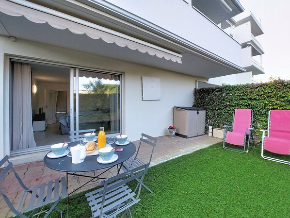 Une terrasse avec une table ronde dressée pour les repas, des chaises d’extérieur et un petit jardin agrémenté d’une haie.