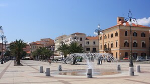A public square with a central fountain, surrounded by palm trees and historic buildings with red-tiled roofs.