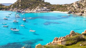 A coastal area with boats on turquoise waters, rocky terrain, and a hill with vegetation.