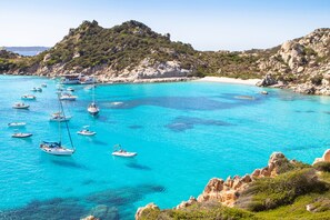A coastal area with boats on turquoise waters, rocky terrain, and a hill with vegetation.