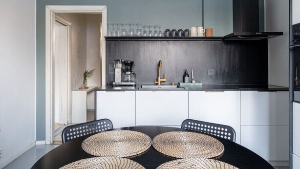 Una cocina moderna con encimera negra, armarios blancos y campana extractora de acero inoxidable. Hay una cafetera y un fregadero con grifo dorado.