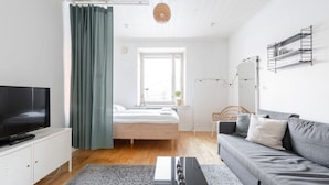 Una sala de estar moderna con un televisor de pantalla plana, un sofá gris, una cama con ropa blanca, una ventana con cortinas y una lámpara colgante.