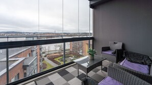 Een balkon met uitzicht op een rivier en gebouwen, een glazen tafel, rieten meubels en een potplant.