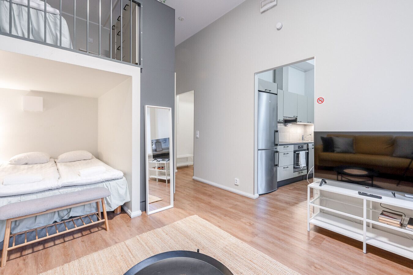 A modern apartment with a loft bedroom, a kitchenette, a living area with a sofa and TV, and a dining table.