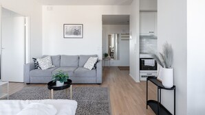 Ein modernes Wohnzimmer mit einem grauen Sofa, einem kleinen Couchtisch mit einer Pflanze und einer Küchenzeile mit Herd und weißen Schränken.