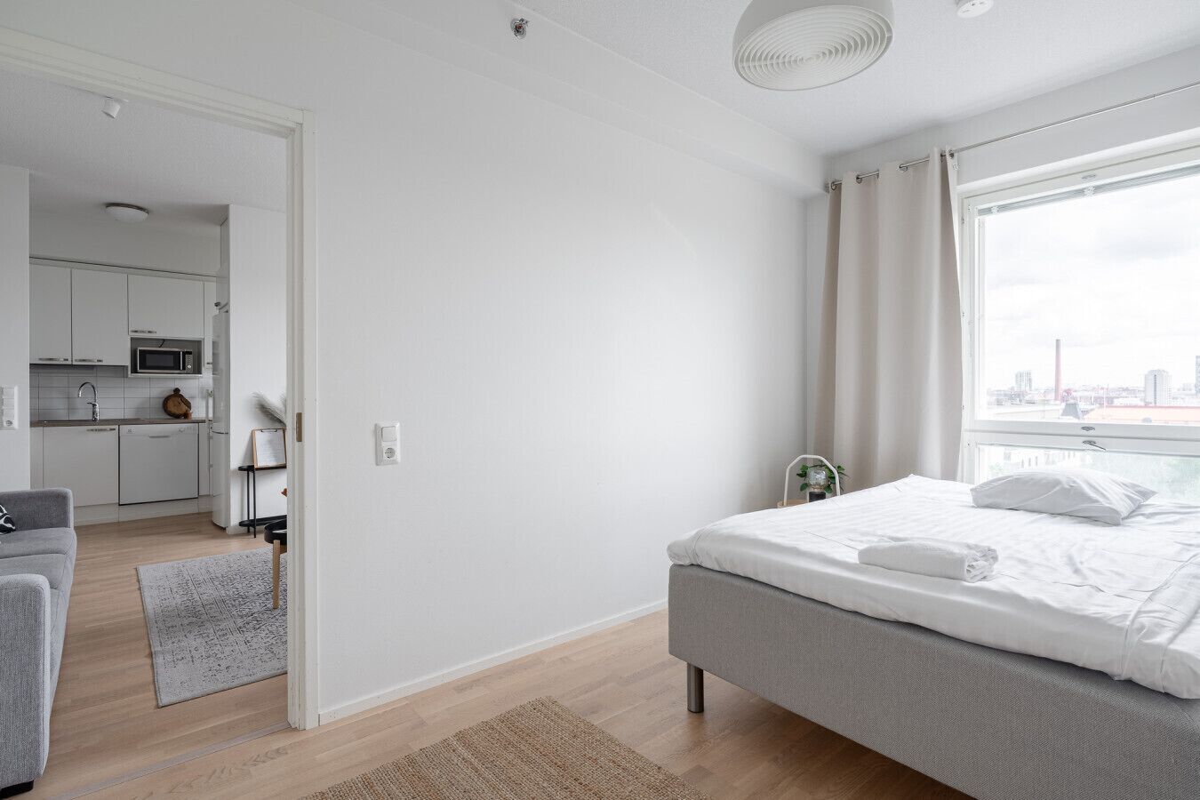 A modern bedroom with a bed, a grey sofa, a small kitchen area, and a view of the city through the window.