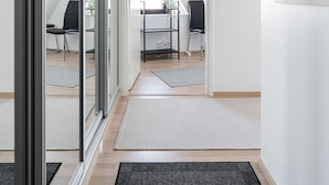 Un pasillo moderno y bien iluminado con una puerta de vidrio que da a un dormitorio tipo loft. Se observan dos sillas de oficina negras, un escritorio con una computadora y una pequeña planta sobre el escritorio. La habitación tiene techo inclinado y vista al exterior.