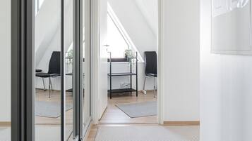 A modern, well-lit hallway with a glass door leading to a loft bedroom. There are two black office chairs, a desk with a computer, and a small plant on the desk. The room has a sloped ceiling and a view of the outdoors.