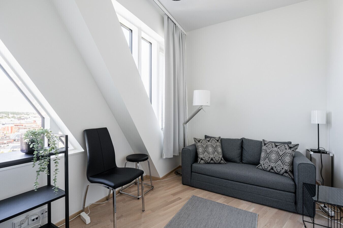 Een moderne woonkamer met een grijze bank, zwarte fauteuil en een klein tafeltje. Er is een raam met gordijnen, een vloerlamp en een plant op een plank.