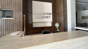 A reception desk with a wooden panel wall, a mounted sign reading "PROSPER RESORT", a small shelf with books, and a desk fan. - Morib Prosper Resort by VP (Banting)