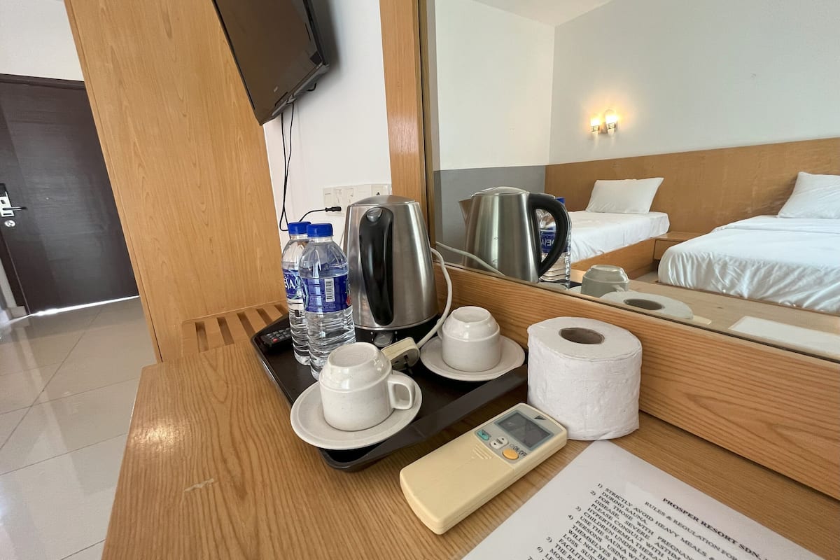 A hotel room with a wooden desk, a coffee maker, two water bottles, a cup, a roll of toilet paper, a remote control, and a mirror reflecting the bed and a wall-mounted light fixture.