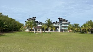A modern building with a large grassy field in front, surrounded by palm trees. - Morib Prosper Resort by VP (Banting)