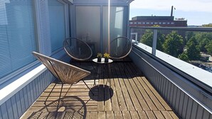 Una terraza de madera con dos sillones de mimbre y una mesita, con vistas a un paisaje urbano que incluye un edificio con la leyenda 'Scandie'.