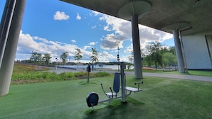 An outdoor exercise area with stationary bikes and a bench, under a concrete structure with a view of a lake and trees.
