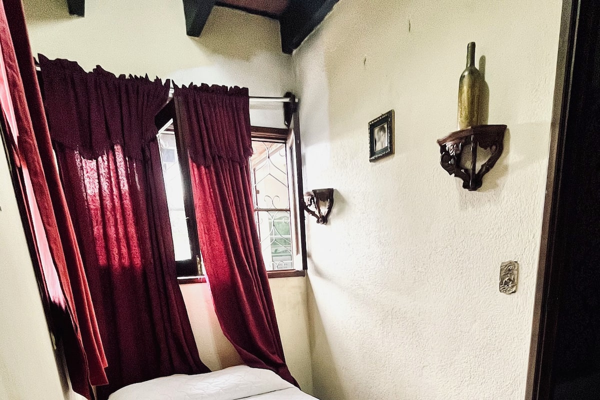 A single bed in a room with a window, red curtains, a wall-mounted light fixture, and a small picture frame.