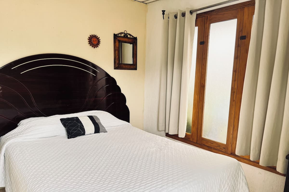 A neatly made bed with a headboard, a window with curtains, and a wall-mounted mirror.
