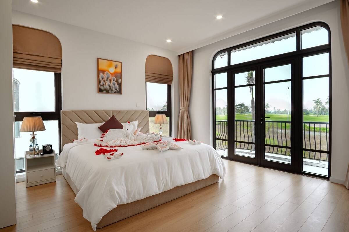 A spacious bedroom with a large bed, a nightstand, a lamp, and a view of a golf course through the windows.