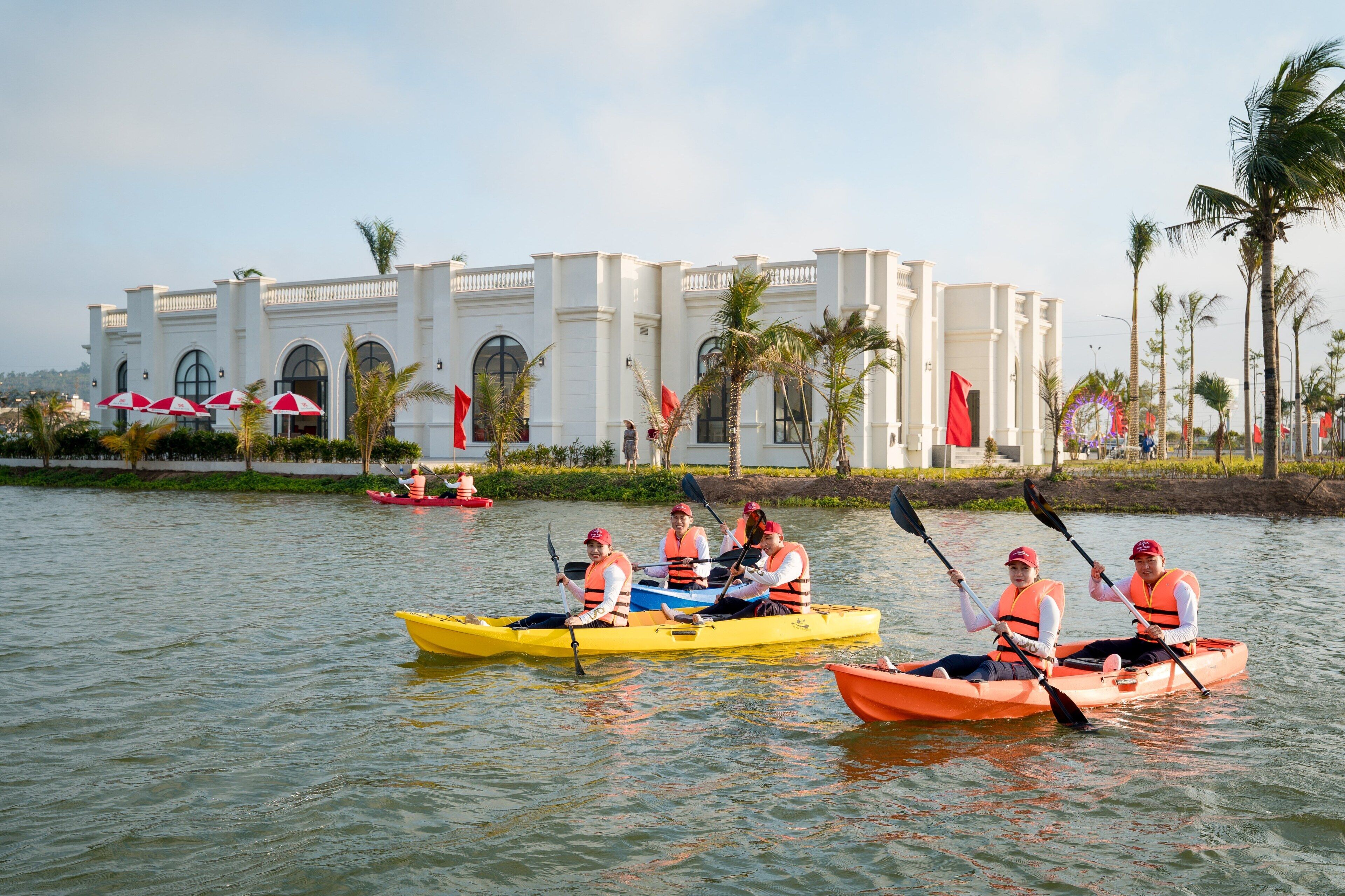 A group of people kayaking on a calm body of water with a white building in the background.