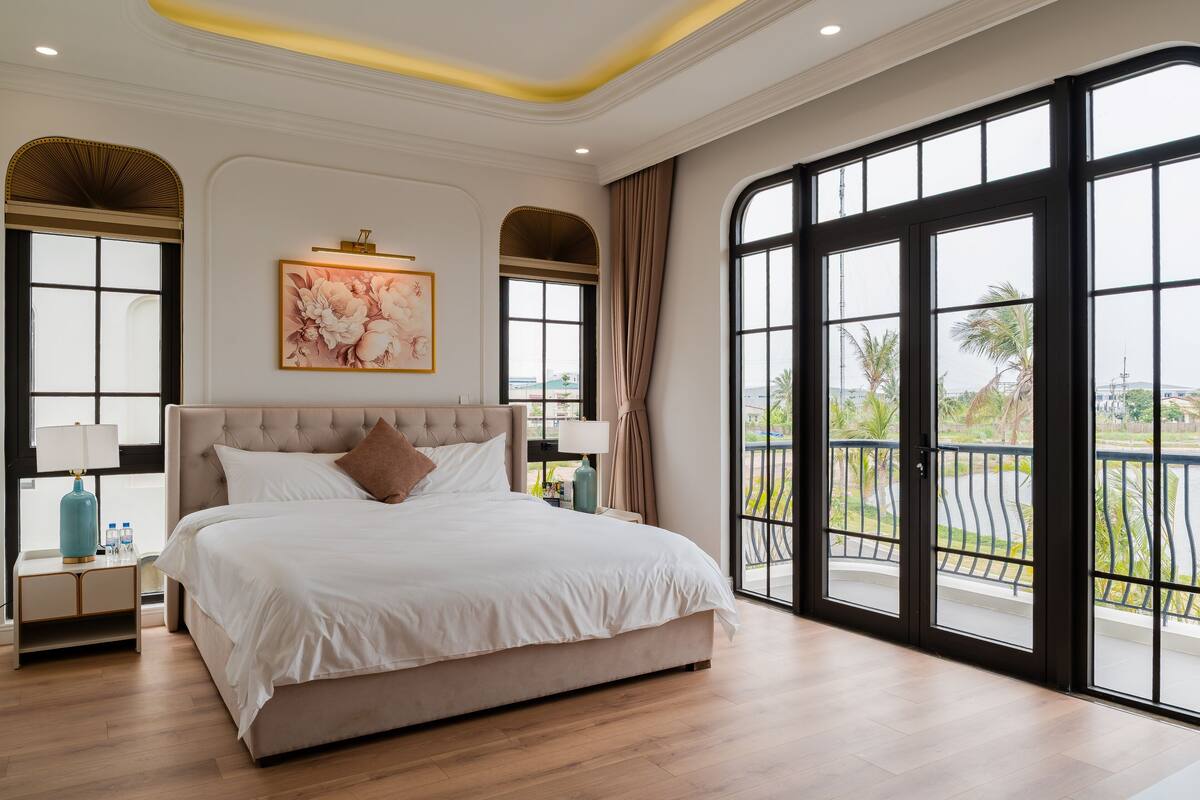 A spacious bedroom with a large bed, a view of the outdoors through large windows, and a balcony with a railing.