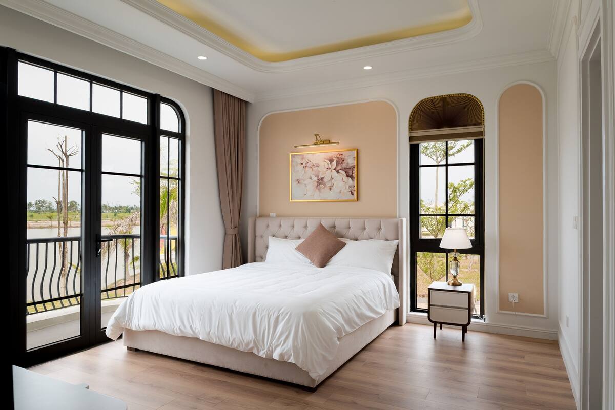 A bedroom with a large bed, a nightstand, a window with a view, and a wall-mounted picture.