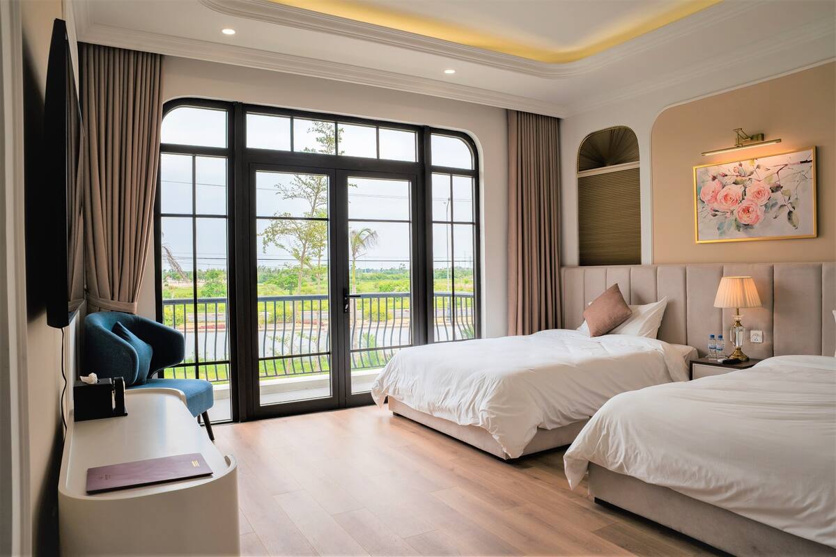 A hotel room with two beds, a desk, a chair, a TV, and a large window with a view of greenery.