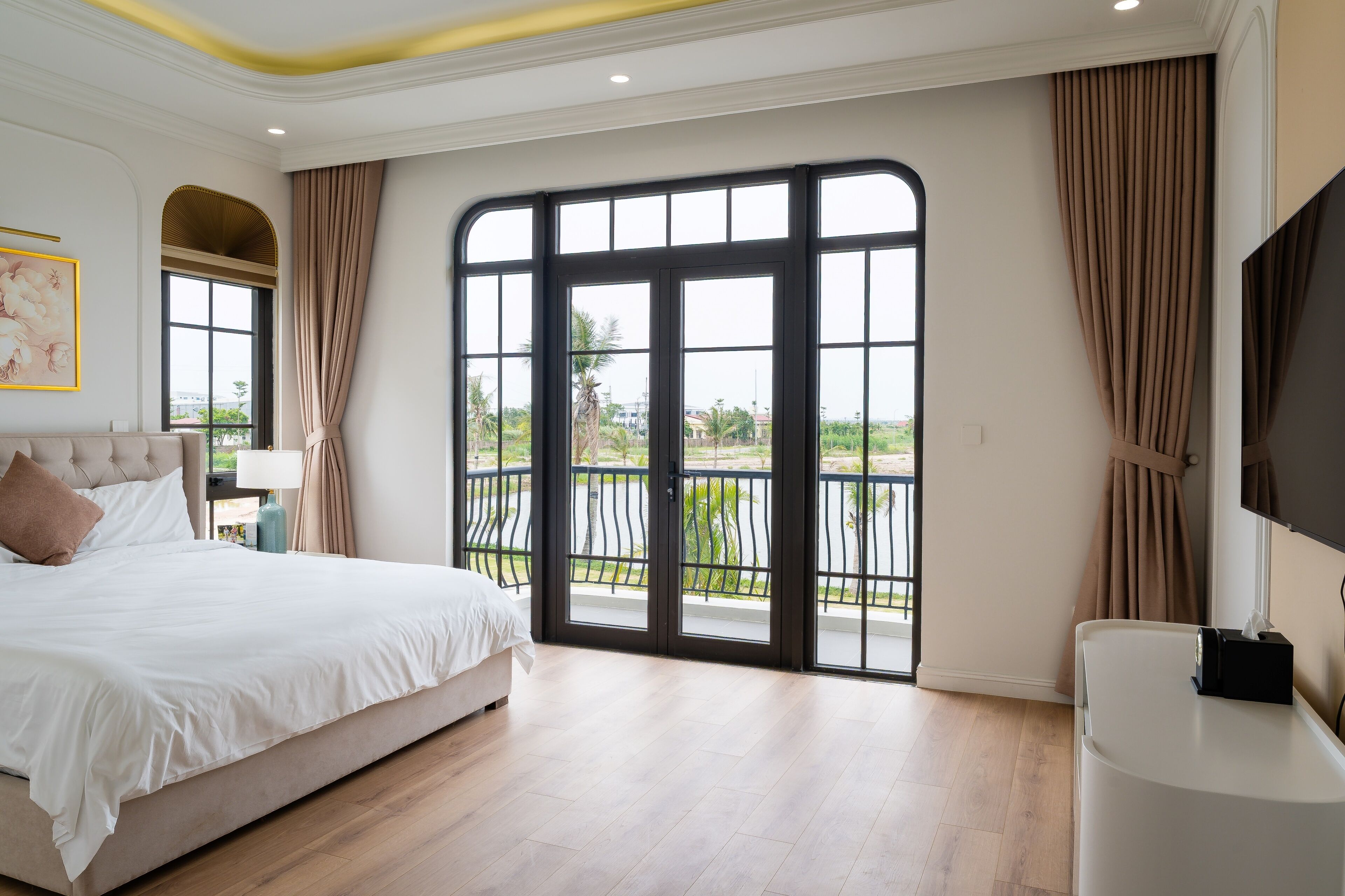 A bedroom with a large bed, a TV, and a balcony with a view of greenery.