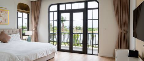 A bedroom with a large bed, a TV, and a balcony with a view of greenery.