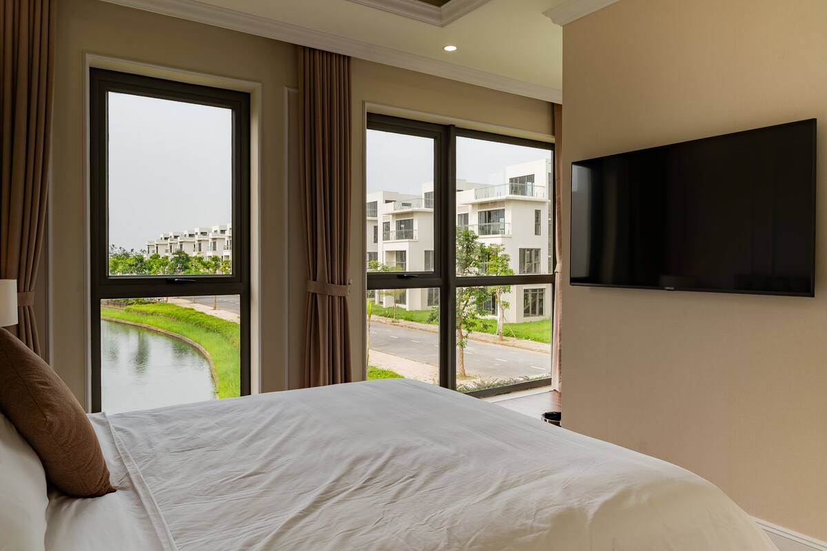 A hotel room with a large bed, a flat-screen TV mounted on the wall, and a view of a canal and buildings through the windows.