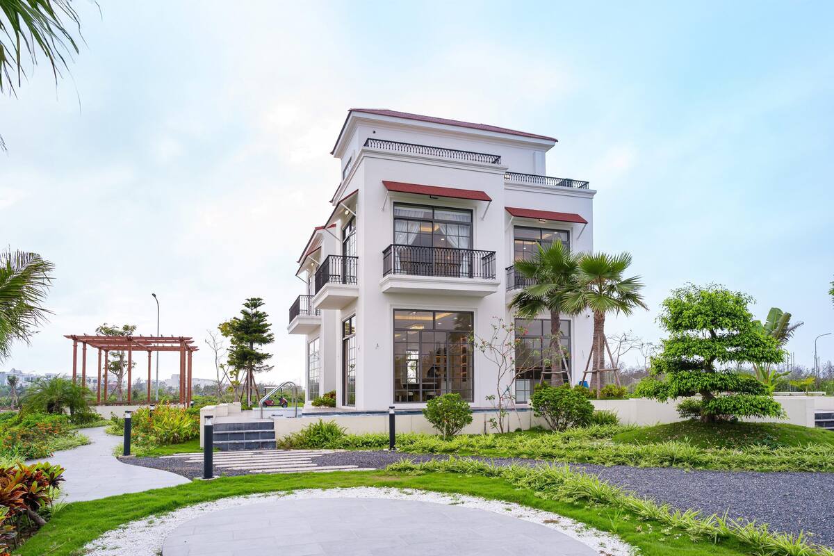 A modern two-story house with a balcony, surrounded by a well-maintained garden with a circular patio.