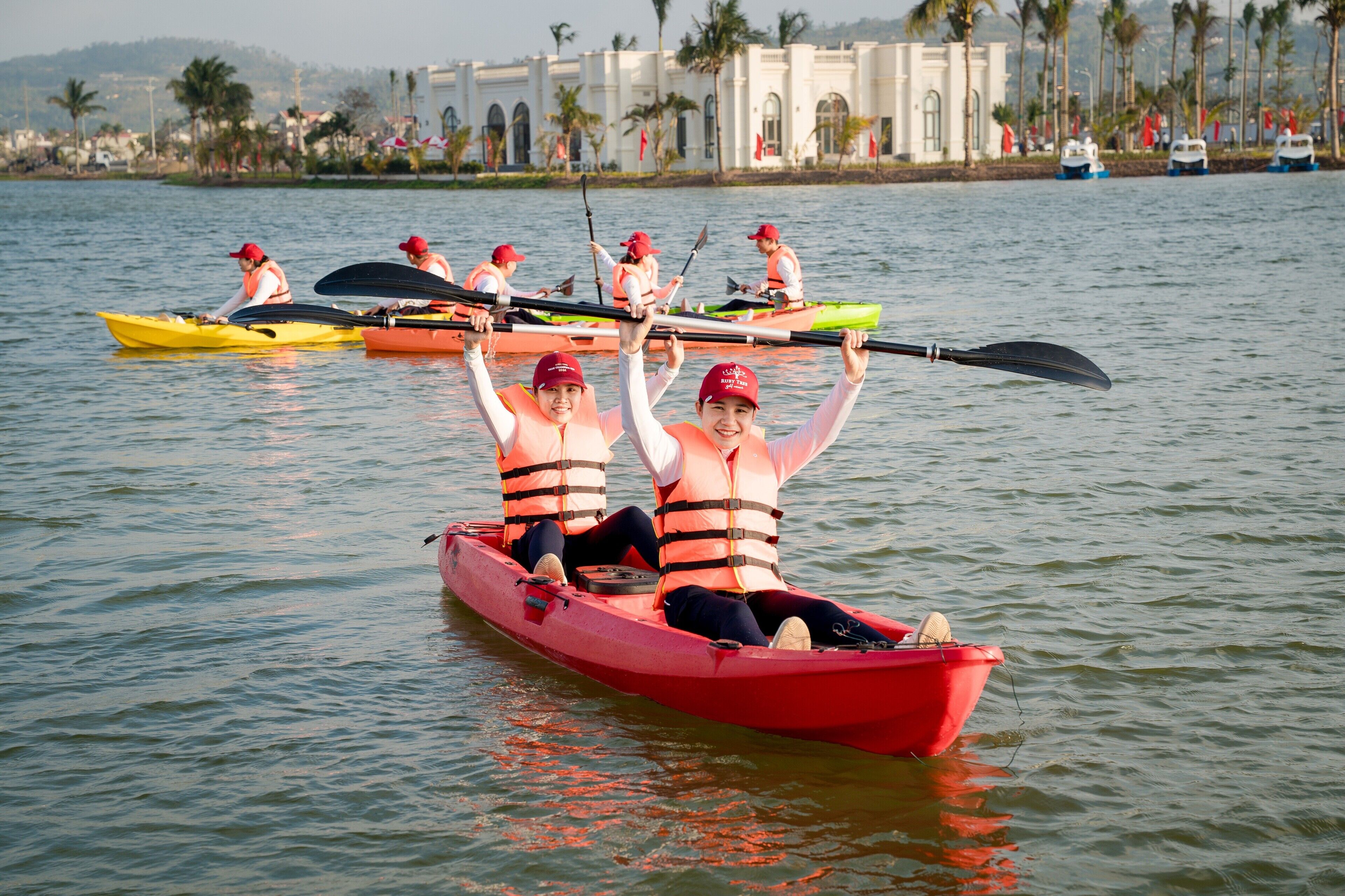 Un groupe de personnes se trouve dans un canoë rouge sur une étendue d’eau calme, avec un bâtiment et des palmiers en arrière-plan.