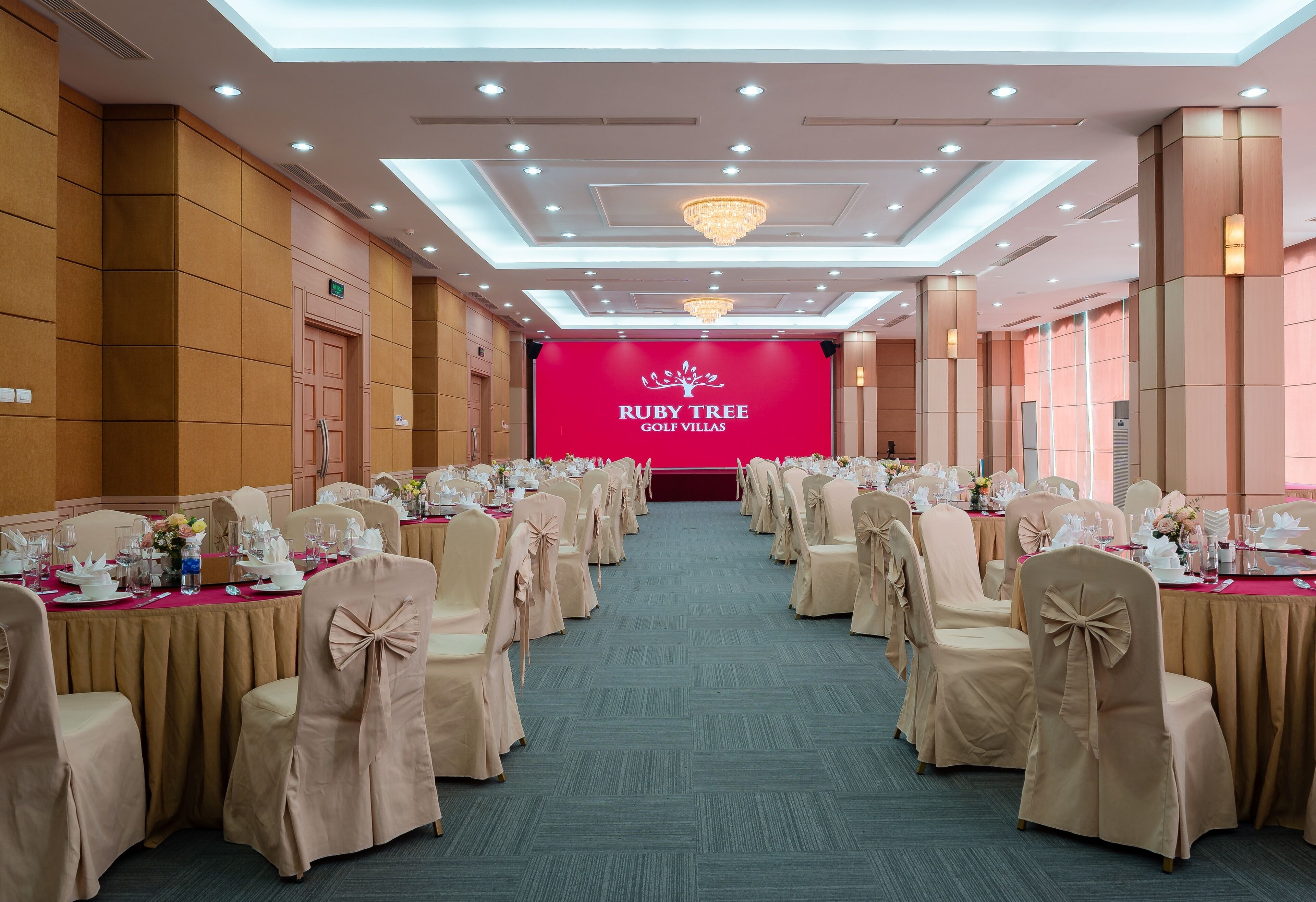 Une salle de banquet avec des rangées de tables dressées pour un événement officiel, une scène ornée d’une banderole portant l’inscription « Ruby Tree Golf Villas », et un lustre.