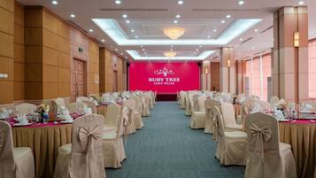 Une salle de banquet avec des rangées de tables dressées pour un événement officiel, une scène ornée d’une banderole portant l’inscription « Ruby Tree Golf Villas », et un lustre.