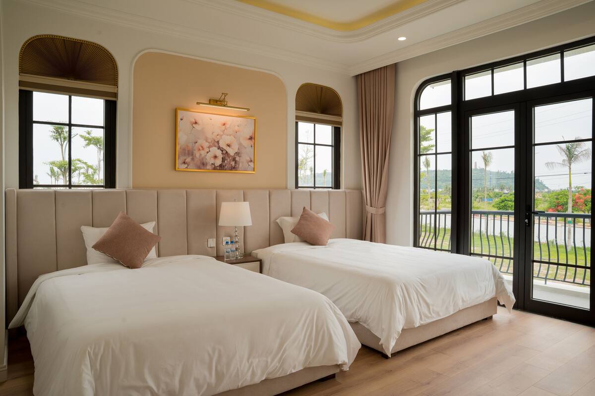 A hotel room with two beds, a large window with a view, a painting on the wall, and a nightstand with a lamp.