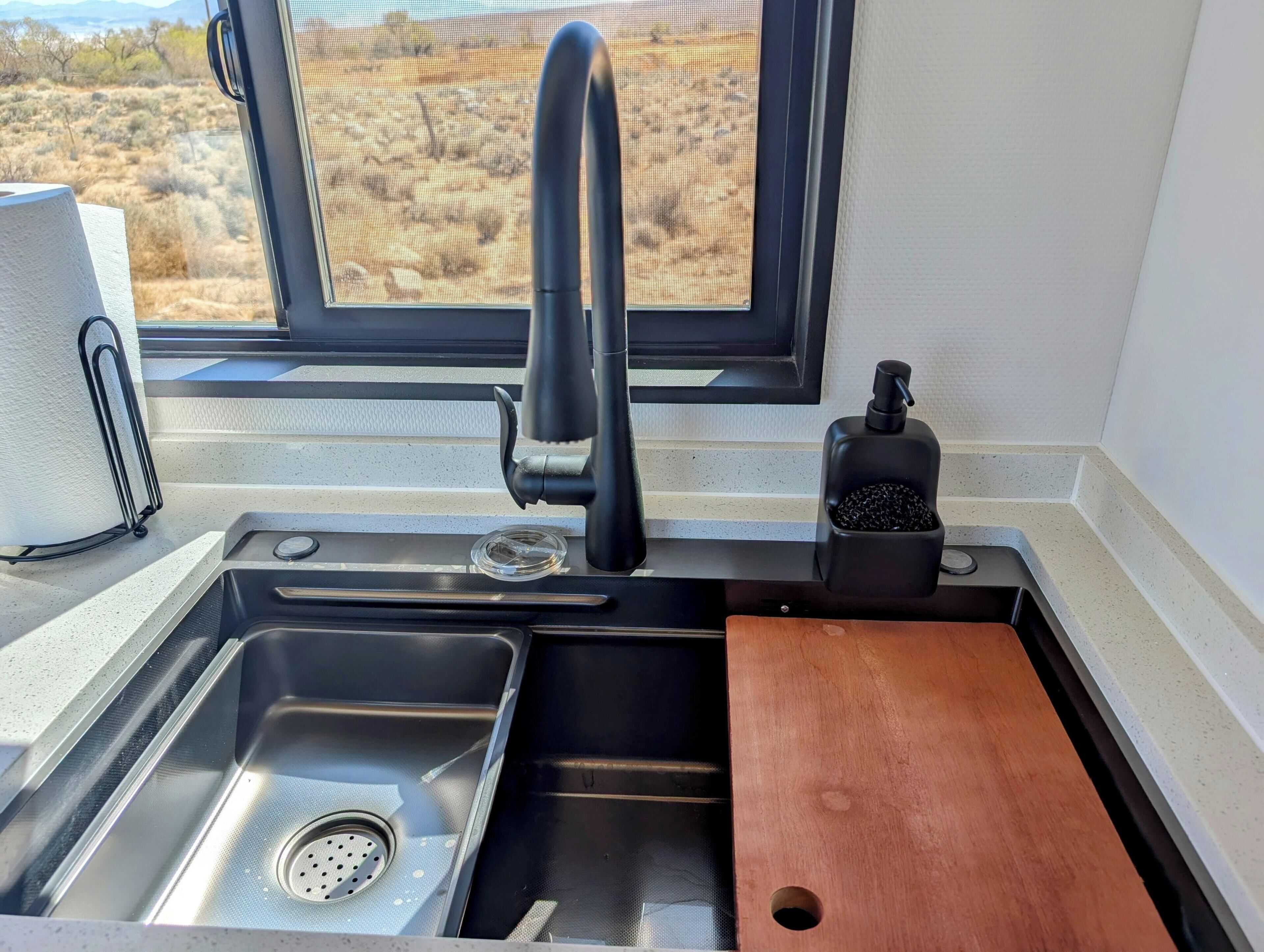 A kitchen with a stainless steel sink, a window with a view of a dry landscape, and a soap dispenser.