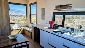 A modern kitchen with a view of a desert landscape, wooden cabinetry, and a dining area with a table and a bowl. - Summit Sanctuary: 360° Views (Lone Pine)