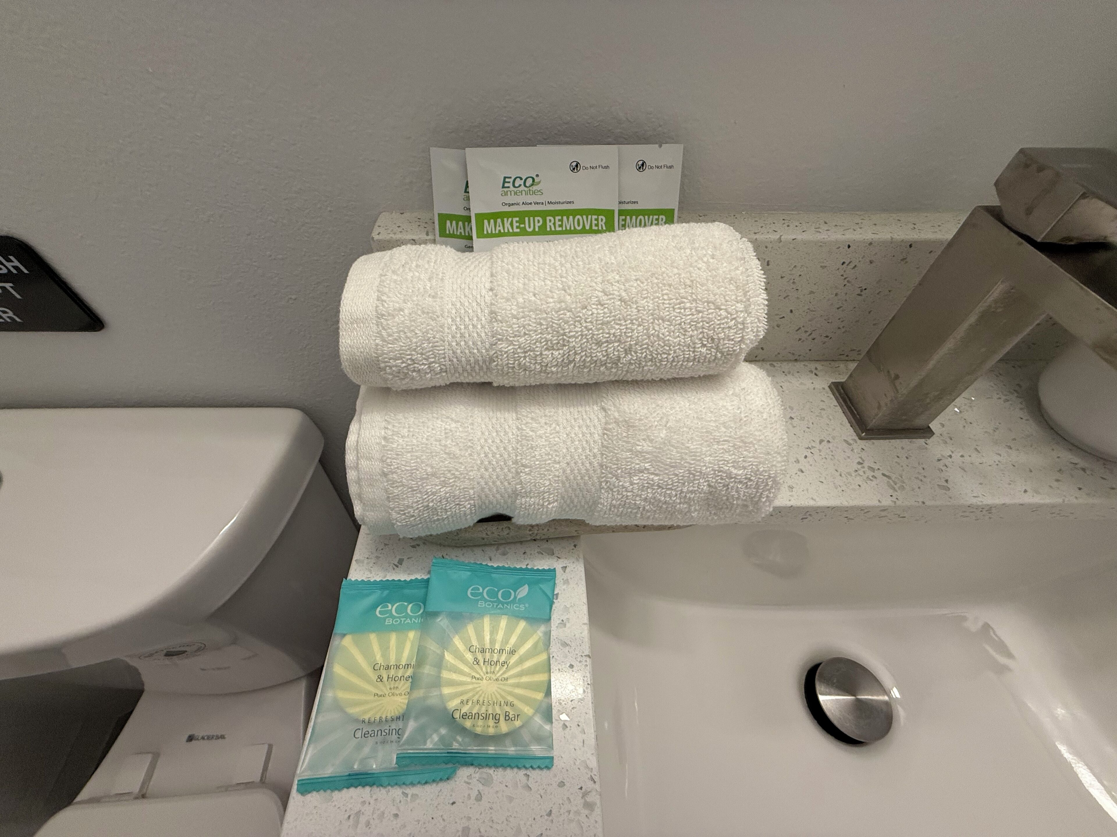 A bathroom with a white sink, a toilet, and a stack of white towels. There are two boxes labeled 'ECO amenities' and 'Make-up remover' on the towel stack.