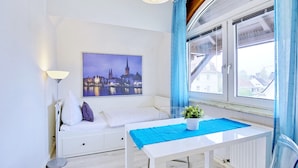 Een kamer met een bed, een tafel met een blauwe tafelkleed, een raam met gordijnen, een lamp en een stadsgezichtschilderij aan de muur.