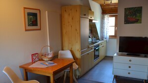 Een kleine keuken met een eethoek, houten kastjes, een televisie en een raam met gordijnen.