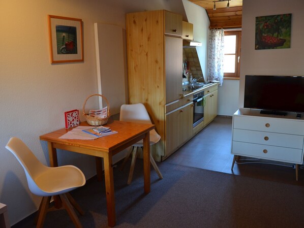 A compact kitchen with a dining area, wooden cabinets, a TV, and a window with curtains. - Studio 'Kirsche' with Shared Garden, Balcony and Wi-Fi (Kressbronn am Bodensee)