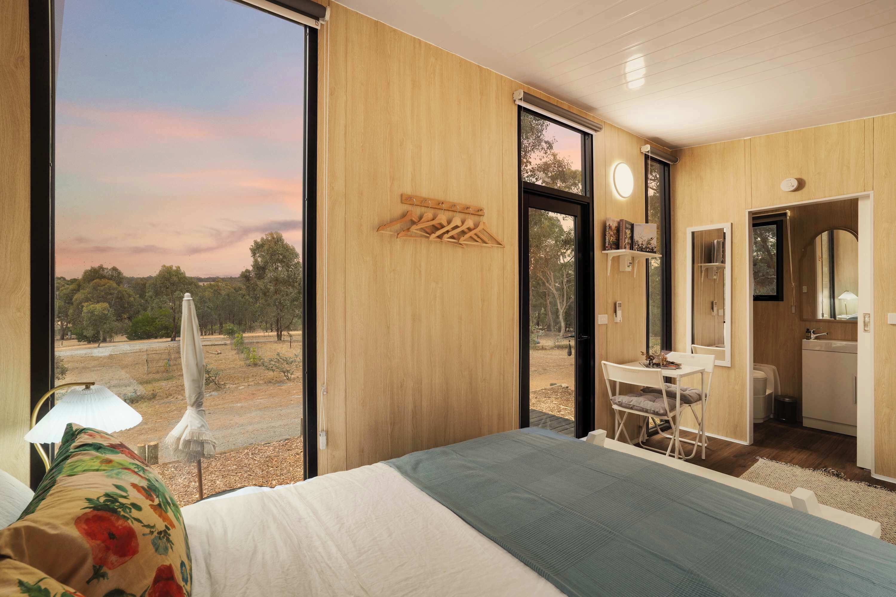 A bedroom with a large window, wooden walls, a bed, a desk, and a bathroom.