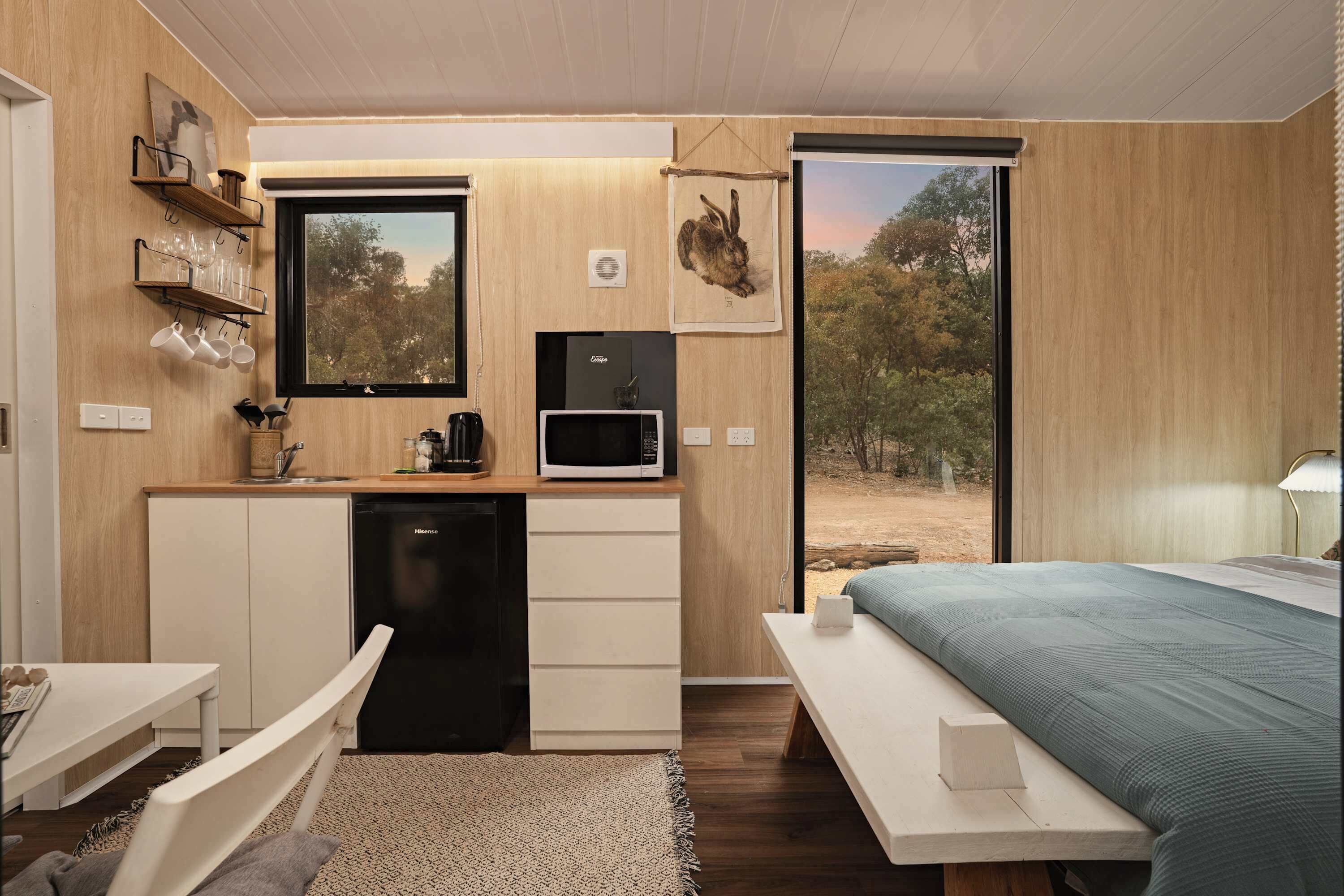 Una habitación compacta con kitchenette, una cama, un ventanal con vistas a árboles y una mesita.