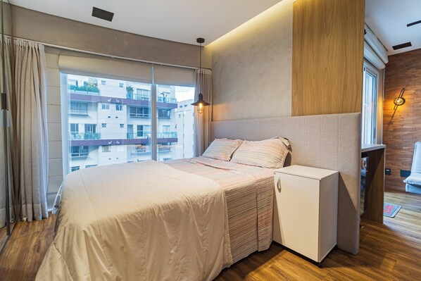 A modern hotel room with a large bed, a small bedside table, a built-in wardrobe, and a view of the city through large windows.