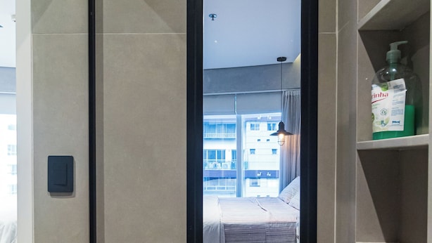 Quarto de hotel com um espelho grande, uma cama, chão de madeira, prateleira com artigos de higiene pessoal e vista para a cidade pela janela.