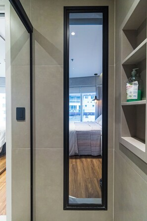 Habitación de hotel con un espejo grande, una cama, piso de madera, estante con artículos de aseo y vista a la ciudad por la ventana.